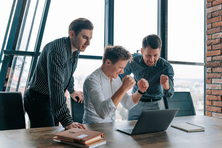 Joyful team of three men celebrate a successful project while working on laptops in a modern office during daylightの写真素材