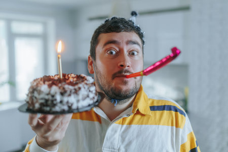 Joyful man wrapped in duvet celebrates birthday in kitchen with cake and whistleの写真素材