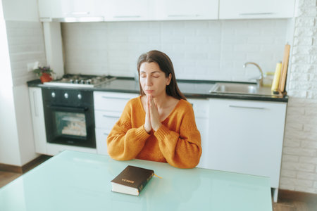 Thoughtful young woman reading and praying in a serene and cozy kitchen.の写真素材