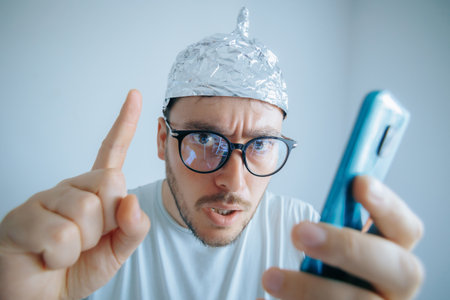 Man in aluminum foil hat expresses disbelief while holding smartphone in roomの写真素材
