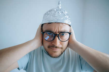Man wearing aluminum foil hat demonstrates conspiracy theory paranoia with humorous expression in indoor settingの写真素材