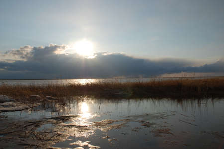 In the evening over the lake, the April sun sets behind the clouds, the sun's rays are reflected in the water.の写真素材