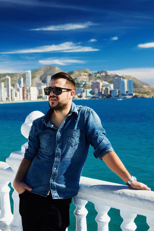 Portrait of trendy guy with beard and sunglasses on a Mediterranean beach, with hand supported to the railing, blurred background. の写真素材