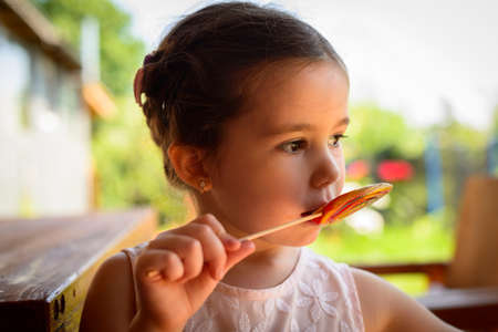 Cute little girl on backyard holding in hand big multicolor spiral lollipop candy, small child wearing pink dress outdoors in summer eating sweet sugar candy, dental health and care conceptの写真素材