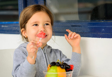 Happy little girl eating fruit salad using fork. Little girl eating healthy snackの写真素材