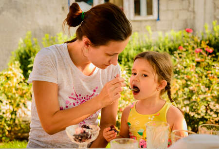 Young beautiful mother feeding her pretty little daughter outdoors in sunlightの写真素材