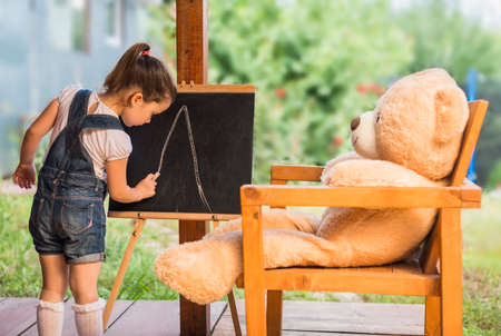 Cute toddler playing teacher role game outdoors. Little child is drawing with pieces of chalk on a blackboard. Happy kid leaning letters and numbers. Children education conceptの写真素材