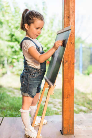 Little child is drawing with pieces of chalk on a blackboard outdoor. Happy kid leaning letters and numbers. Children education conceptの写真素材