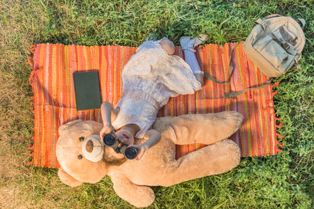 Adorable little girl with binoculars looking to the sky lying down with teddy bear on the picnic blanket outdoor in a sunny summer day. Looking for the summer vacationの写真素材