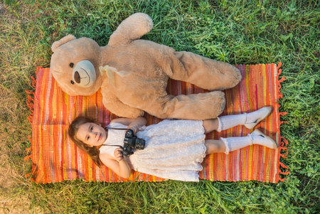 Young little girl photographer with old vintage film photo camera laying down on picnic blanket with teddy bear outdoor in a sunny summer day. Children's play. Art or creativity concept.の写真素材