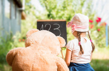 Cute toddler playing teacher role game with her Teddy bear friend outdoors. Happy kid leaning numbers. Children education conceptの写真素材