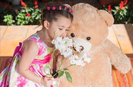 Outdoor portrait of expressive little girl with white flowers bouquet hugging huge plush bear.の写真素材