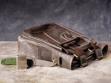 Green folding EDC knife and silver lighter near a leather business casual bagの写真素材