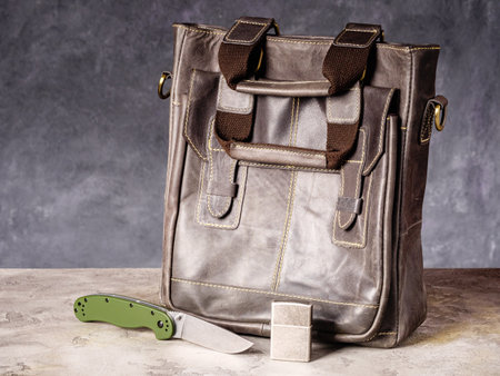 Green folding EDC knife and vintage lighter near a leather business casual bagの写真素材