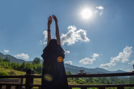 the girl stretches, looking at the morning sun and mountains. Next to her, on the railing of the balcony, is a cup of coffee.の写真素材