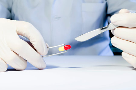 Hands in medical rubber gloves holding a scalpel and tweezers with red and yellow capsule.