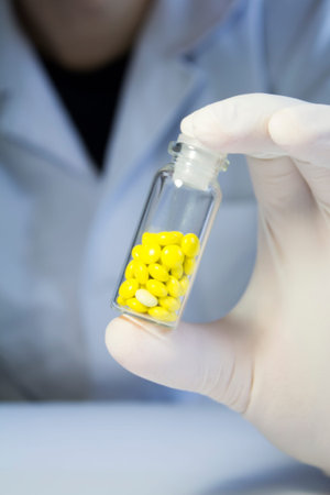 Close-up hand in medical rubber glove holding glass jar with yellow pills.の写真素材