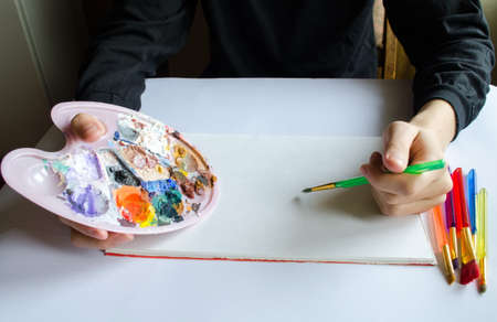 The left hand draws a brush with blue paint on paper in an album with several colorful brushes near it.の写真素材