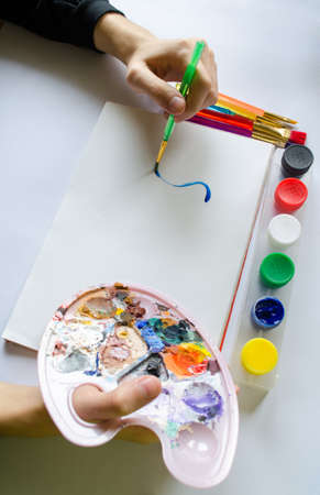 The left hand draws a brush with blue paint on paper in an album with several colorful brushes near it.の写真素材