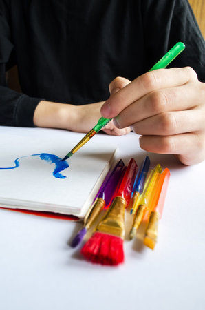 The left hand draws a brush with blue paint on paper in an album with several colorful brushes near it.の写真素材