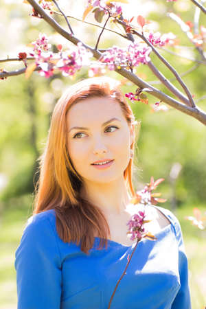 Beautiful young red-haired girl in blue dress among spring flowers in blossom garden.の写真素材