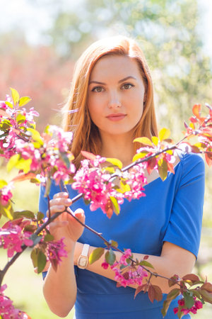 Beautiful young red-haired girl in blue dress among spring flowers in blossom garden.