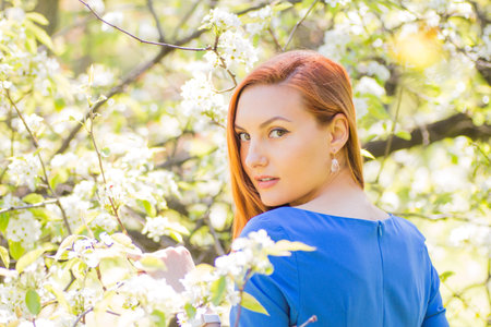 Beautiful young red-haired girl in blue dress among spring flowers in blossom apple tree garden.の写真素材
