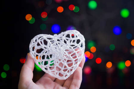Knitted white heart in hand with garland lights on colorful bokeh background.の写真素材