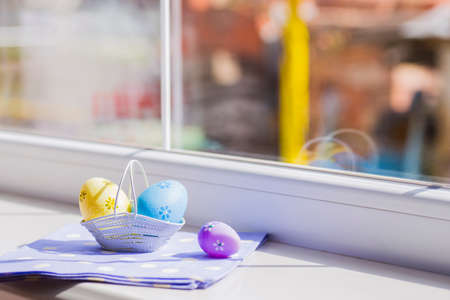 Colorful easter eggs in white basket on blue fabric near window.の写真素材