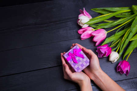 Gift box in hands with bouquet of pink tulips on black wooden backgroundの写真素材