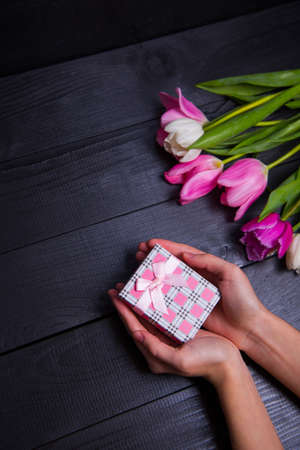 Gift box in hands with bouquet of pink tulips on black wooden backgroundの写真素材