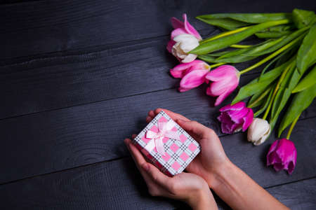 Gift box in hands with bouquet of pink tulips on black wooden backgroundの写真素材