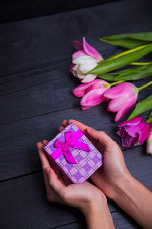 Gift box in hands with bouquet of pink tulips on black wooden backgroundの写真素材