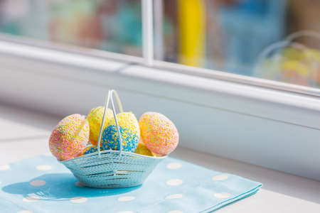 Colorful easter eggs in white basket on blue fabric near window.の写真素材