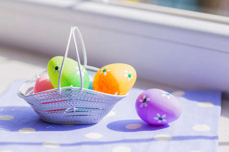 Colorful easter eggs in white basket on blue fabric near window.の写真素材