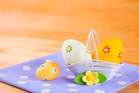 Colorful yellow and orange Easter eggs in white basket with yellow flower on spotted fabric on wooden backgroundの写真素材
