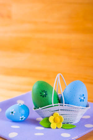 Colorful blue and green Easter eggs in white basket with yellow flower on spotted fabric on wooden backgroundの写真素材