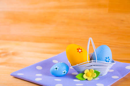 Colorful blue and orange Easter eggs in white basket with yellow flower on spotted fabric on wooden backgroundの写真素材