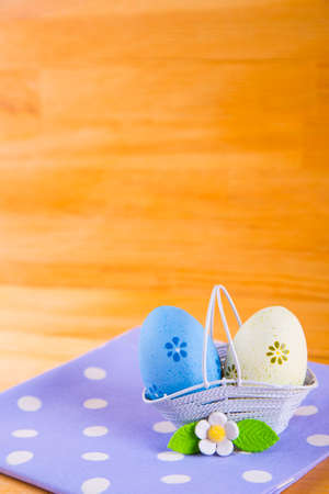 Two colorful blue and yellow Easter eggs in white basket with white flower on spotted fabric on wooden backgroundの写真素材