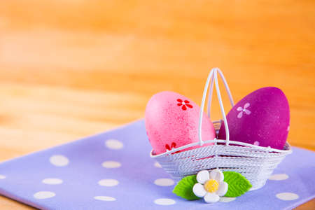 Two colorful pink and crimson Easter eggs in white basket with white flower on spotted fabric on wooden backgroundの写真素材