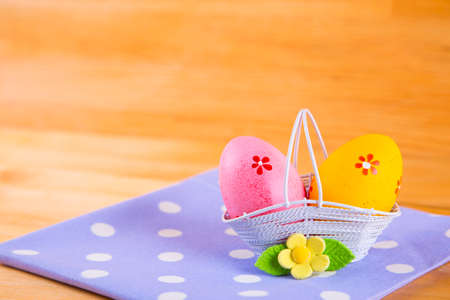 Two colorful pink and orange Easter eggs in white basket with yellow flower on spotted fabric on wooden backgroundの写真素材