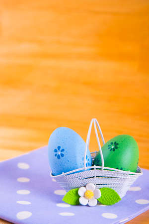Two colorful blue and green Easter eggs in white basket with white flower on spotted fabric on wooden backgroundの写真素材