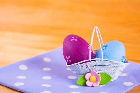 Two colorful blue and crimson Easter eggs in white basket with pink flower on spotted fabric on wooden backgroundの写真素材