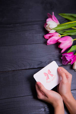 Beautiful bouquet of tender pink and white tulips with female hands holding greeting card on black wooden background. Spring romantic composition.の写真素材