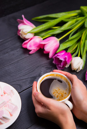 Beautiful bouquet of tender pink and white tulips with female hands holding cup of coffee and saucer with marshmallows on black wooden background. Spring romantic composition.の写真素材