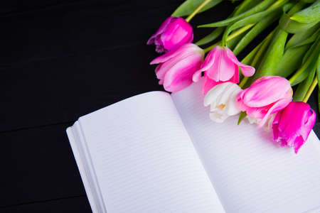 Beautiful bouquet of tender pink and white tulips and open notebook on black wooden background. Spring romantic composition.の写真素材