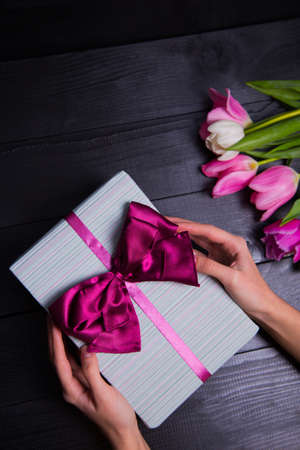 Beautiful bouquet of tender pink and white tulips with female hands holding gift box on black wooden background. Spring romantic composition.の写真素材