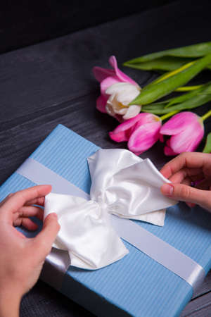 Beautiful bouquet of tender pink and white tulips with female hands packing and wrapping gift box on black wooden background. Spring romantic composition.の写真素材
