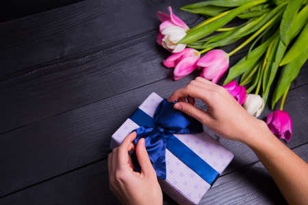 Beautiful bouquet of tender pink and white tulips with female hands packing and wrapping gift box on black wooden background. Spring romantic composition.の写真素材