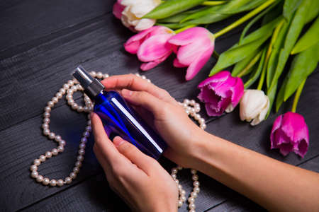 Beautiful bouquet of tender pink and white tulips with female hands holding blue bottle of perfume on black wooden background with pearl necklace. Spring romantic composition.の写真素材